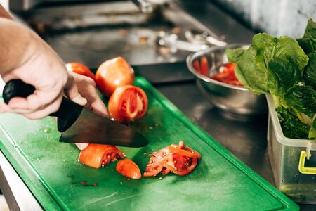 partial view of cook cutting tomatoes on chopping board in kitchenの写真素材