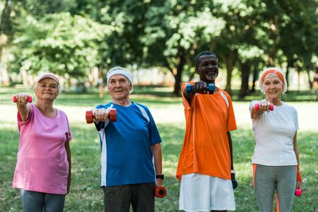smiling multicultural pensioners exercising with dumbbells in parkの写真素材
