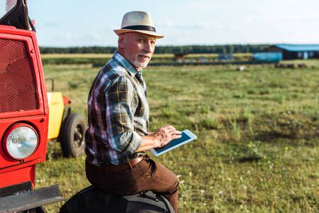 happy farmer in straw hat using digital tablet near red tractor
