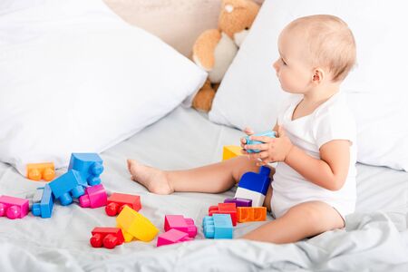 Barefoot little child in white clothes sitting on bed with pillows near colorful toys