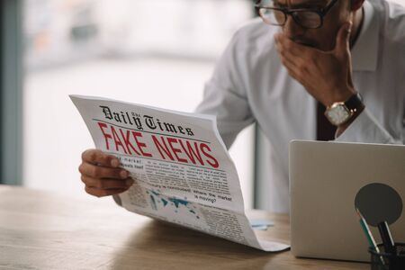 shocked businessman in glasses covering mouth while reading newspaper with fake newsの写真素材