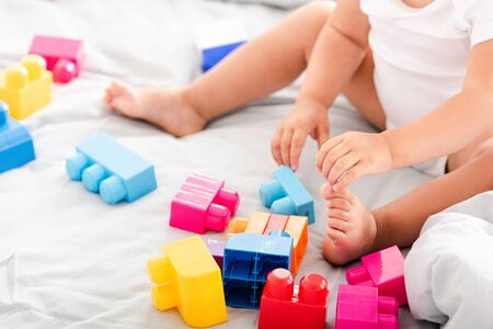 Partial view of barefoot baby in white clothes sitting on bed and playing with bright construction