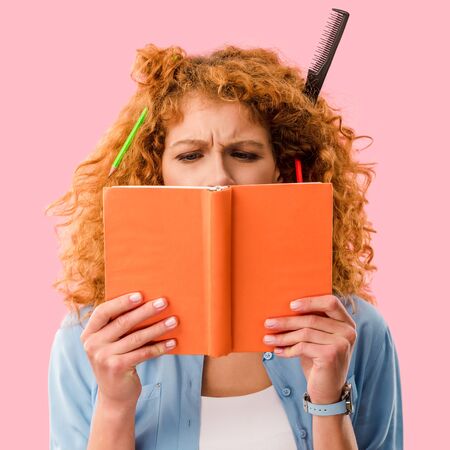 focused student with pencils in hair holding book isolated on pink
