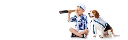 panoramic shot of smiling boy in sailor suit looking through spyglass and beagle dog on white