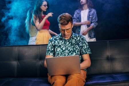 handsome man in sunglasses sitting on couch and using laptop in nightclubの写真素材