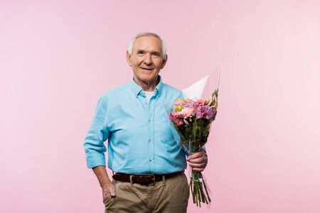 cheerful senior man standing with hand in pocket and holding bouquet on pinkの写真素材