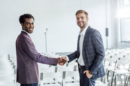 two multiethnic colleagues in formal wear shaking hands in conference hallの写真素材