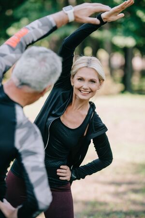 smiling sportswoman warming up in park near mature sportsman