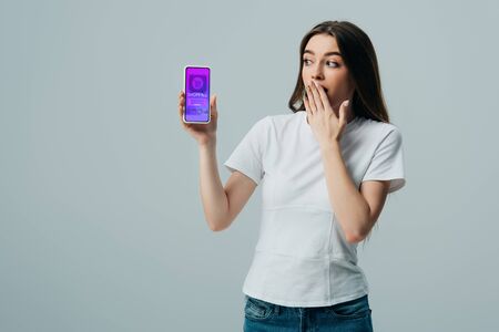 shocked beautiful girl in white t-shirt showing smartphone with online shopping app isolated on grey