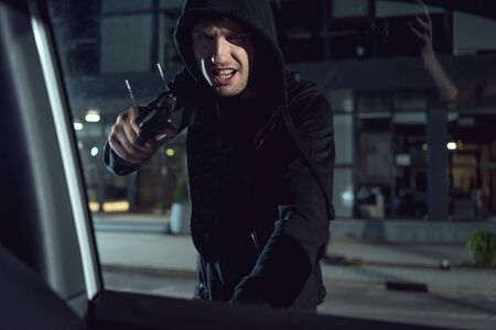 aggressive thief in black hoodie pointing gun at car windowの写真素材