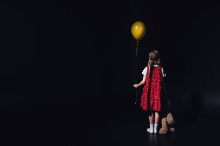 back view of depressed child with yellow balloon and teddy bear isolated on blackの写真素材