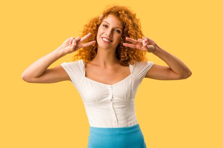 cheerful woman with red hair showing peace symbols isolated on yellowの写真素材