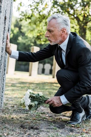 upset man with grey hair putting flowers near tomb in cemeteryの写真素材