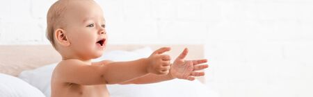 Panoramic shot of adorable little child opening mouth and raising hands forward in white roomの写真素材