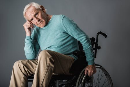 upset retired man sitting in wheelchair and looking at camera on greyの写真素材