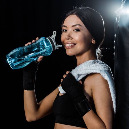cheerful girl holding sport bottle and smiling isolated on black