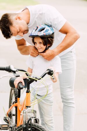 father putting helmet on son while boy standing near bicycle and looking awayの写真素材