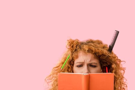concentrated redhead student with pencils in hair holding book isolated on pinkの写真素材