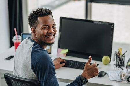 cheerful african american programmer looking at camera and showing thumb up while sitting near computersの写真素材