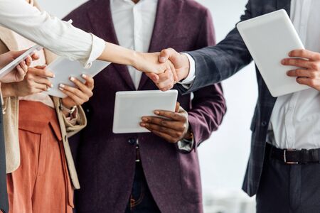 cropped view of four multiethnic colleagues holding digital tablets and shaking hands
