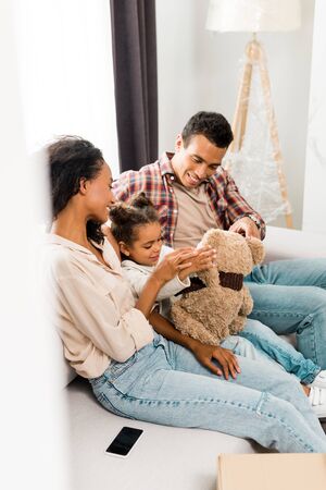 selective focus of african american family sitting on sofa and smiling while daughter playing with teddy bear and parents looking at kidの写真素材