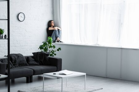 depressed young woman sitting on window sill at home and looking away