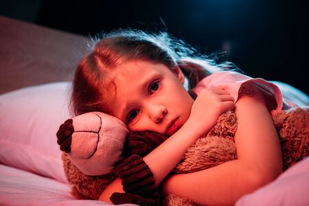 scared child hugging teddy bear while lying in bed and looking at camera isolated on blackの写真素材