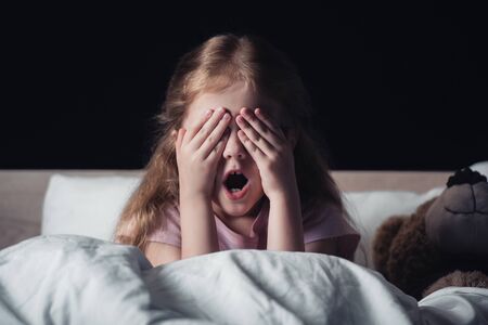 frightened kid screaming and covering eyes with hands while sitting on bedding near teddy bear isolated on blackの写真素材