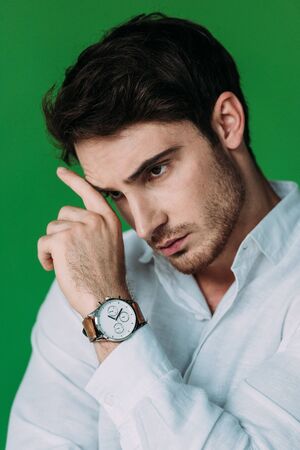 serious pensive man in white shirt and wristwatch touching face with hand isolated on green