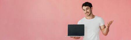 panoramic shot of smiling young man in white t-shirt showing laptop with blank screen isolated on...
