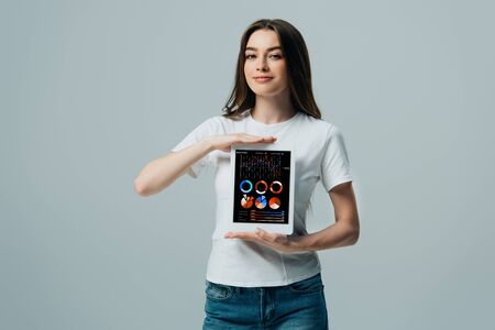 smiling beautiful girl in white t-shirt showing digital tablet with financial app isolated on greyの写真素材