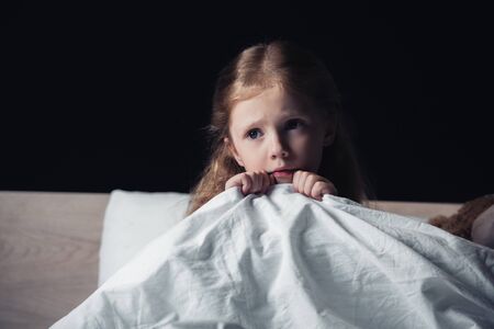 scared child looking away while sitting on bedding in dark bedroom isolated on blackの写真素材