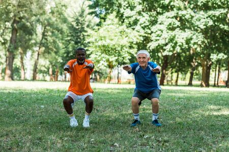 happy senior and multicultural med doing sit ups on grass near treesの写真素材