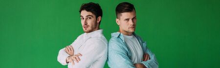 panoramic shot of two serious friends standing with crossed arms and looking at camera isolated on...