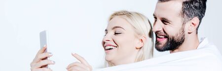 panoramic shot of man and beautiful girl covered in blanket using smartphone on white