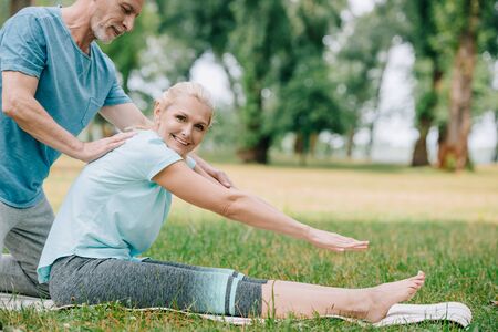 handsome mature man helping smiling woman practicing yoga in parkの写真素材