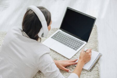 back view of woman with headphones lying on floor, holding pen and and looking at screen of laptopの写真素材