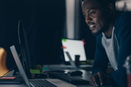 young african american programmer smiling while looking at computer monitor at night in officeの写真素材