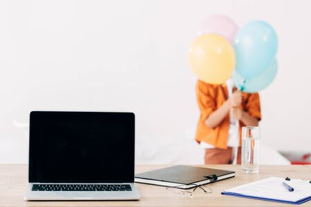 selective focus of kid holding colorful balloons and laptop with blank screen on foregroundの写真素材