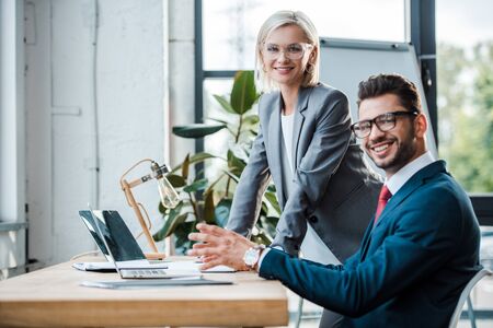 cheerful coworkers in glasses looking at camera near laptops in officeの写真素材