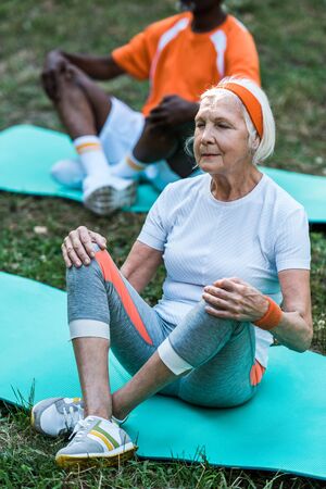 selective focus of senior woman in sportswear and african american man sitting on fitness matsの写真素材