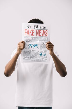 african american man covering face while reading newspaper with fake news lettering isolated on whiteの写真素材