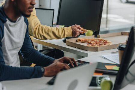 cropped view of programmer holding piece of pizza near african american colleagueの写真素材