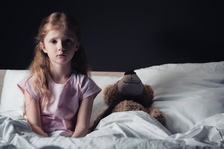 frightened child sitting on bedding near teddy bear and looking at camera isolated on blackの写真素材