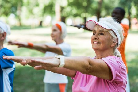 selective focus of retired woman doing exercise near multicultural pensioners in park