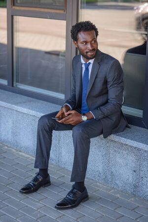 smiling african american businessman looking away while sitting on parapet and holding smartphoneの写真素材