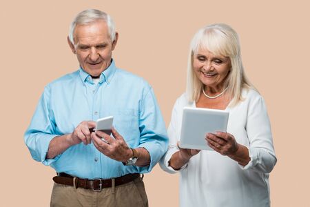 happy retired man using smartphone near wife with digital tablet isolated on beigeの写真素材
