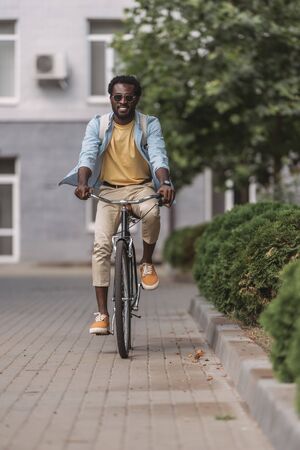 handsome, stylish african american man smiling while riding bicycle along streetの写真素材