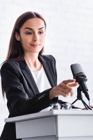 smiling lecturer touching microphone while standing on podium tribune in conference hallの写真素材