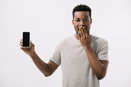 shocked african american man covering face and holding smartphone with blank screen isolated on whiteの写真素材